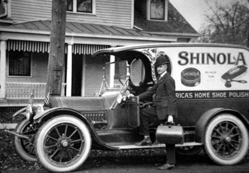 Black and white photo of a man in uniform seated in an early 20th-century Shinola shoe polish delivery truck parked in front of a house.